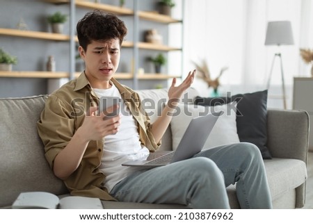 Confused asian man looking at phone screen, sitting on couch with computer on lap in living room, upset frustrated male reading bad news on message, having problem with device or Internet connection Royalty-Free Stock Photo #2103876962