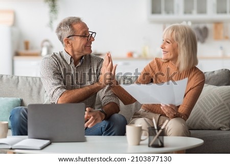 Joyful Mature Couple Holding Papers And Giving High-Five Celebrating Financial Success Sitting On Couch In Front Of Laptop Reading Bills At Home. Successful Budgeting And Household Concept Royalty-Free Stock Photo #2103877130