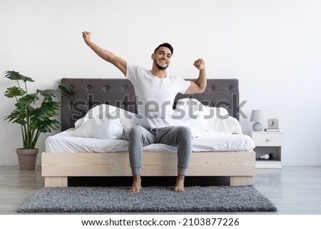 Handsome Happy Young Arab Guy Waking Up In The Morning, Sitting On Bed And Stretching After Good Sleep, Smiling Millennial Middle Eastern Man Having Good Mood, Enjoying Start Of New Day, Copy Space Royalty-Free Stock Photo #2103877226