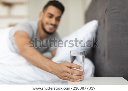 Smiling Middle Eastern Man Taking Glass Of Water From Bedside Table In Bedroom, Thirsty Young Arab Guy Reaching Hand To Healthy Drink After Waking Up In The Morning, Selective Focus With Free Space Royalty-Free Stock Photo #2103877319