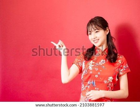 Smiling asian young woman in traditinal red cheongsam dress pointing hand to empty space on red background.