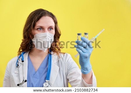 A doctor in a blue uniform on a yellow background holds two injection syringes. Face of a nurse in a medical hat and mask, close-up of a coronavirus vaccine