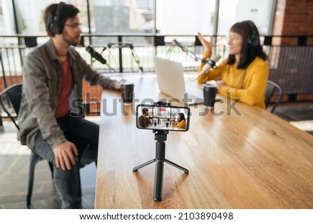 Two podcasters, man and woman talking while making live video podcast in studio, using microphones and headphones. Woman host streaming live video with man guest. Selective focus on smartphone screen