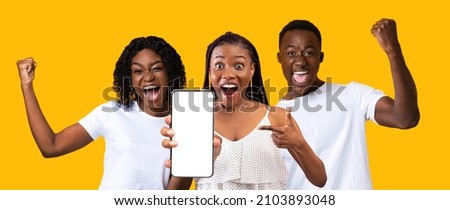 Happy millennial friends black guy and two ladies in white showing smartphone with empty screen, having fun, screaming and raising hands up on yellow studio background, panorama, mockup