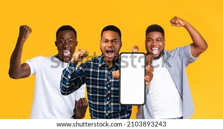 Emotional black guys three friends having fun together, gambling online, showing smartphone with empty screen on yellow studio background, screaming and raising fists up, mockup, panorama