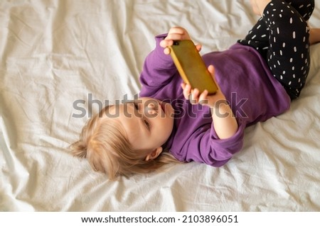Sweet little girl gen Z using online app on smartphone, browsing internet, playing videogames, taking selfie, making call, watching cartoons, resting on bed in bedroom at home.
