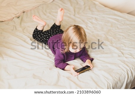 Sweet little girl gen Z using online app on smartphone, browsing internet, playing videogames, taking selfie, making call, watching cartoons, resting on bed in bedroom at home.