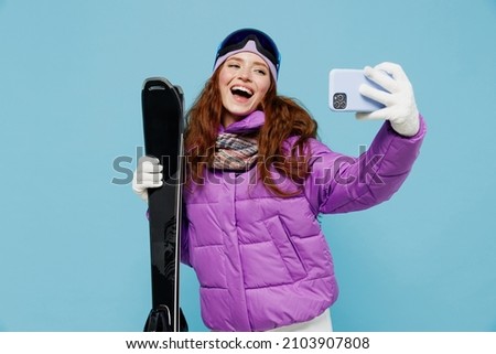 Skier cool woman in warm purple windbreaker jacket goggles mask spend extreme weekend in mountains doing selfie shot on mobile cell phone post photo hold ski isolated on plain blue background studio