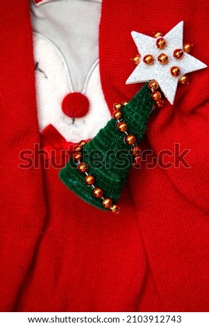 Vertical photo. Small Christmas tree with silver star on top and golden garland on red background. New year composition. Red X-mas sweater with funny face. Postcard picture. Winter party set. 