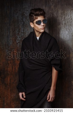 Dark hair handsome young boy with glasses  in wearing in black mantle and black jeans stand and posing on the background black wall indoor