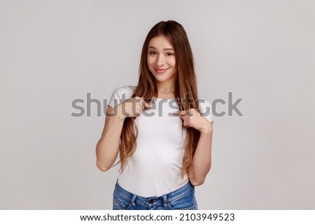 This is me. Portrait of happy young woman pointing herself and smiling, being glad about own success, proud of achievement, wearing white T-shirt. Indoor studio shot isolated on gray background. Royalty-Free Stock Photo #2103949523