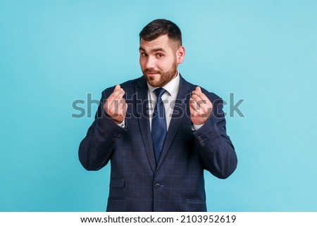 Portrait of handsome bearded young man wearing official style suit standing and looking at camera with money or italian gesture with hand. Indoor studio shot isolated on blue background.