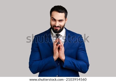 Cunning bearded man clasping hands and planning evil tricky prank or scheming, cheating with sly smile, wearing official style suit. Indoor studio shot isolated on gray background. Royalty-Free Stock Photo #2103954560