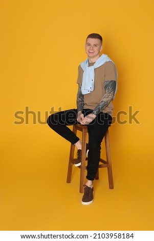 Smiling young man with tattoos sitting on stool against yellow background