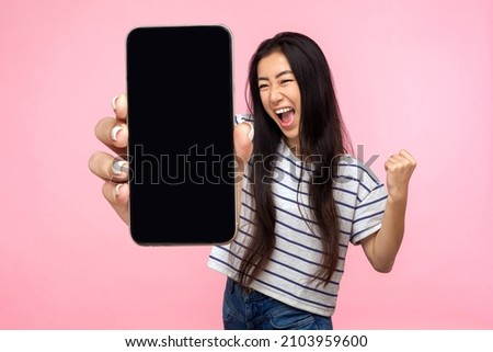 Portrait of euphoric happy joyful girl with long hair screaming with happiness showing mobile advertisement mockup area and celebrating her victory. indoor studio shot isolated on pink background