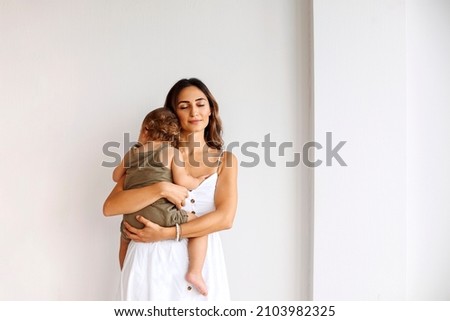 Portrait of motherhood. Young beautiful loving mother wearing white dress holding cute baby boy while standing isolated over white wall. Mom cuddling little son and expressing unconditional love