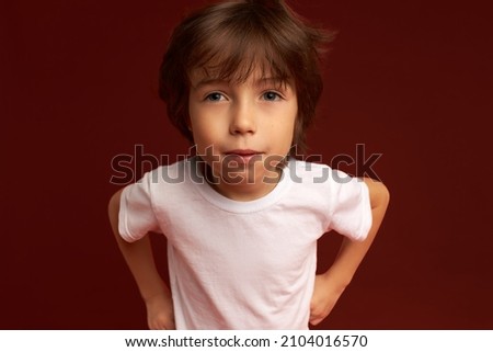 Close-up picture of suspicious casually dressed little man holding hands on waist, looking cool and bossy, bending forward to camera. Children and their emotions, feelings, body language