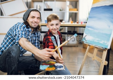 Handsome caucasian man and cute boy looking at camera while holding in hands color palette and paint brushes. Portrait of teacher and student at art school.