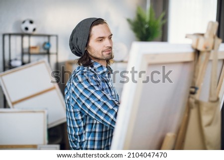Handsome caucasian man drawing with brush on canvas at his painting studio. Young male artist wearing stylish grey hat and checkered shirt.