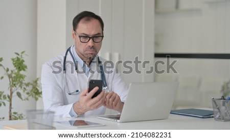 Middle Aged Doctor using Smartphone while Working on Laptop in Clinic