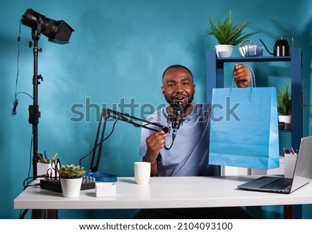Vlogger holding boom arm microphone presenting giveaway blue paper gift bag in vlogging studio with professional live setup. Influencer showing sponsor gift to fans siting at desk with laptop.