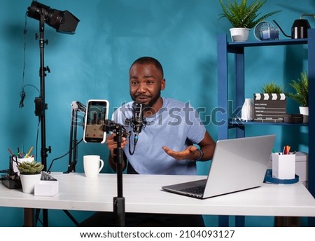 Influencer sitting down at desk with laptop in vlogging studio smiling in front of recording smartphone holding microphone. Content creator interacting with fans looking at live video podcast setup.