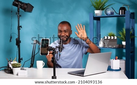 Smiling vlogger waving hello in front of recording smartphone sitting at desk with professional microphone. Content creator doing greeting hand gesture looking at live video podcast setup.