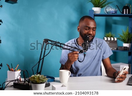 African american vlogger reading live text messages from fans looking at smartphone screen in recording desk in vlogging studio. Influencer having questions and answers session using microphone.