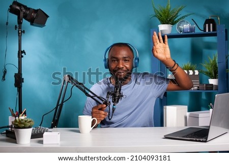Vlogger wearing wirelss headphones waving hello at audience during online live show sitting at desk holding microphone. Content creator doing greeting hand gesture in recording studio.