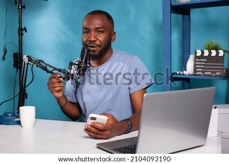 Content creator talking close into professional microphone holding smartphone sitting in front of laptop at podcast recording desk. Influencer answering messages recieved on social media mobile app.
