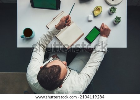 Businessman sitting in a chair at the office table writing in a copybook and using his phone. Top view. Smart office. Business concept.