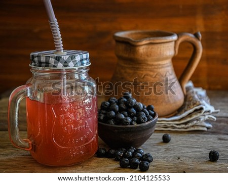 On the wooden kitchen table, there is a ceramic bowl with black rowan berries, a glass cup with black rowan kvass on a kitchen napkin, and an earthen jug. Village life. Healthy drinks. Ethnoscience. Royalty-Free Stock Photo #2104125533