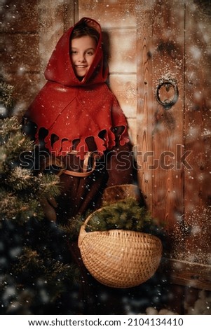 Once upon a Christmas in Medieval times. A cute joyful little girl in medieval clothes stands by a wooden house on a snowy night with a basket of fir branches brought from the forest. 