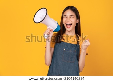 Smile Asian woman wear apron and holding megaphone with fists clenched celebrating victory expressing success on yellow background. Royalty-Free Stock Photo #2104160234