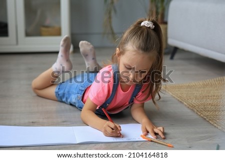 Smilling happy girl lying on warm floor with a toy elephant near to her enjoying creative activity, drawing pencils coloring pictures in albums