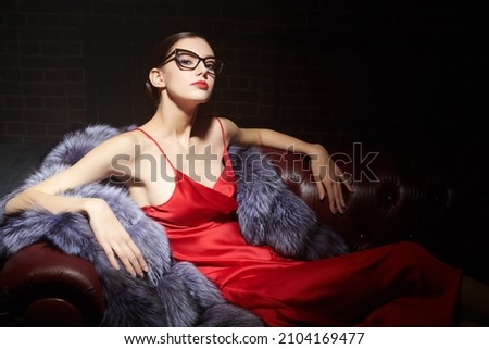 Luxury style in clothes and accessories. Portrait of an elegant brunette girl posing in fashionable glasses, red satin dress and silver fox fur coat on a leather sofa. Fashion shot.