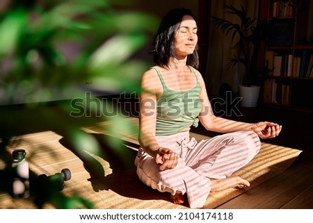 Woman practicing yoga and meditation at home sitting in lotus pose on yoga mat, relaxed with closed eyes. Mindful meditation concept. Wellbeing. Royalty-Free Stock Photo #2104174118