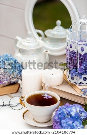 A visual for content. Still life in vintage style. A mug with a drink, an old book, a cage, candles and hydrangea flowers in the garden on a white wooden table. The concept of a tea ceremony.