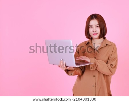 Portrait studio cutout shot Asian young pretty short hair female student model in long sleeve brown shirt stand smiling holding typing browsing surfing learning via laptop computer on pink background.