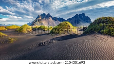 Dramatic afternoon view of Stokksnes cape with Vestrahorn (Batman Mountain) on background. Amazing summer landscape of black sand dunes with green grasson the top. Travel to Iceland. Royalty-Free Stock Photo #2104301693