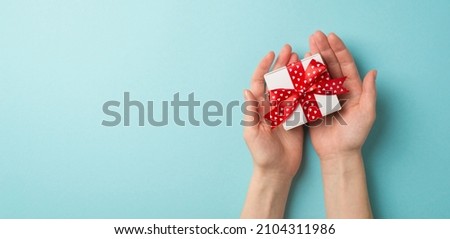 First person top view photo of valentine's day decorations female hands giving small white giftbox with red ribbon bow on isolated pastel blue background with blank space