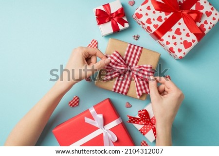 First person top view photo of valentine's day decor present boxes decorative hearts girl's hands tying checkered ribbon bow on craft paper giftbox on isolated pastel blue background with copyspace