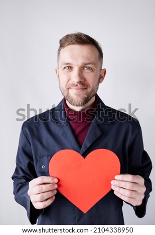 Half length portrait of young bearded hipster man standing by the white wall holding red heart shape. Worker with paper red heart. Romance, relationships concept. Valentine's Day. High quality image