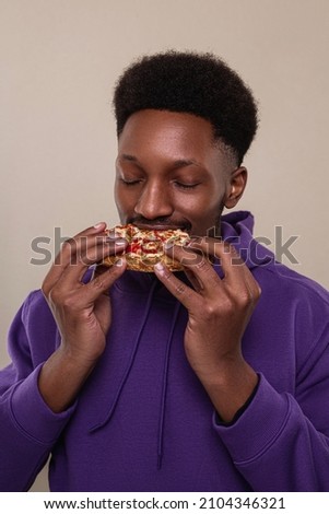 Hungry black man bites very big piece of pizza, has appetite, enjoys eating fast food in office Royalty-Free Stock Photo #2104346321