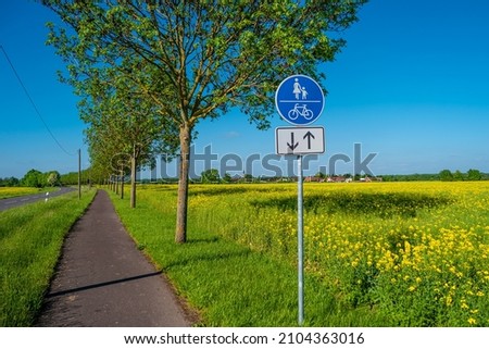 View over beautiful farm landscape with rapeseed blossom field and a cycling and hiking lane or path in Germany, Spring, at sunny day and blue sky, with a signpost for bicycles and people