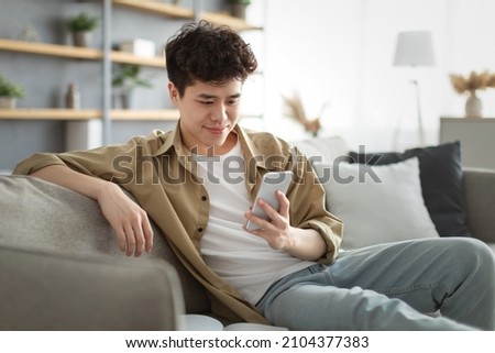 Enjoying Weekend. Handsome Relaxed Asian Man Using Smartphone While Sitting Leaning On Couch At Home, Happy Guy Resting In Living Room, Browsing Internet Or Messaging With Friends, Selective Focus Royalty-Free Stock Photo #2104377383