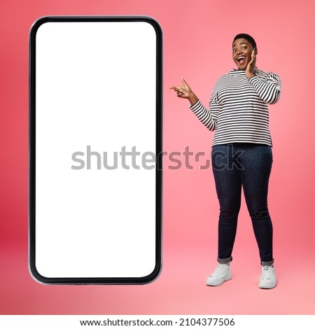Amazed African American Woman Pointing Finger At Huge Smartphone With Empty Screen Advertising Mobile App On Pink Studio Background. Excited Lady Standing Near Big Phone Touchscreen. Square, Mockup