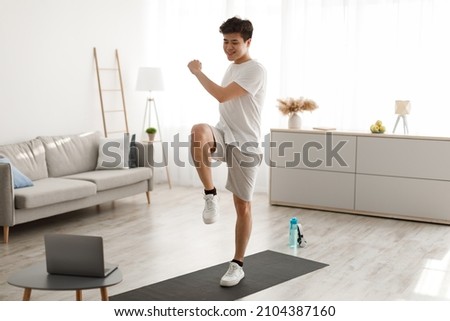 Online Workout Concept. Cheerful Smiling Asian Guy Training In Living Room, Doing Twist Knees To Elbow Exercise. Happy Man Standing And Lifting Leg Up, Using Laptop Watching Tutorial Lookign At Screen Royalty-Free Stock Photo #2104387160