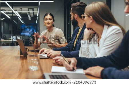 Young businesswoman leading a discussion during a meeting with her colleagues. Group of diverse businesspeople working together in a modern workplace. Business colleagues collaborating on a project. Royalty-Free Stock Photo #2104396973