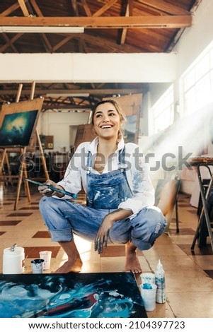 Inspired young artist looking away with a smile in her art studio. Happy female painter contemplating new creative ideas for her art project. Imaginative young woman painting on a canvas on the floor. Royalty-Free Stock Photo #2104397723